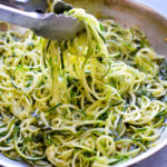 Tongs holding raw zucchini noodles in a bowl.