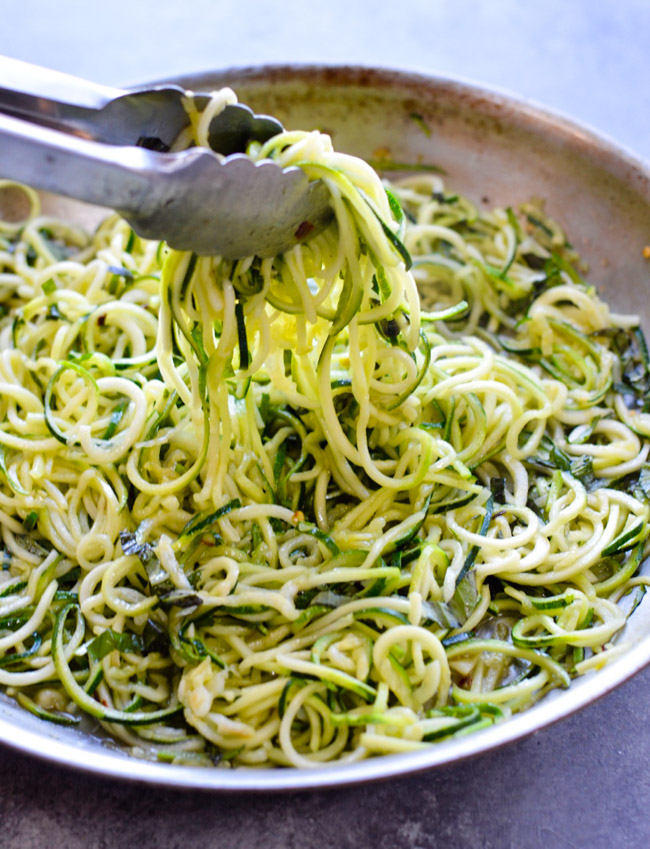 Tongs holding raw zucchini noodles in a bowl.