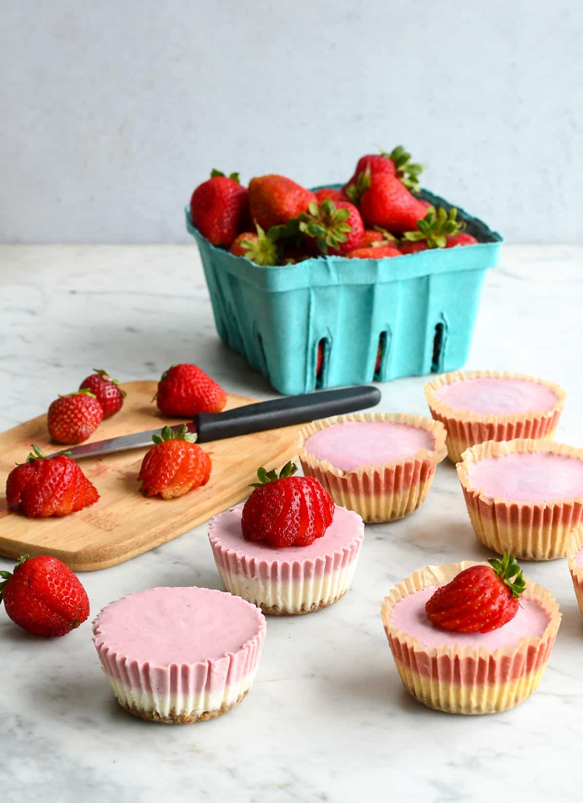 Mini Strawberry Cheesecakes with c cutting board making strawberry fans and a basket of fresh strawberries