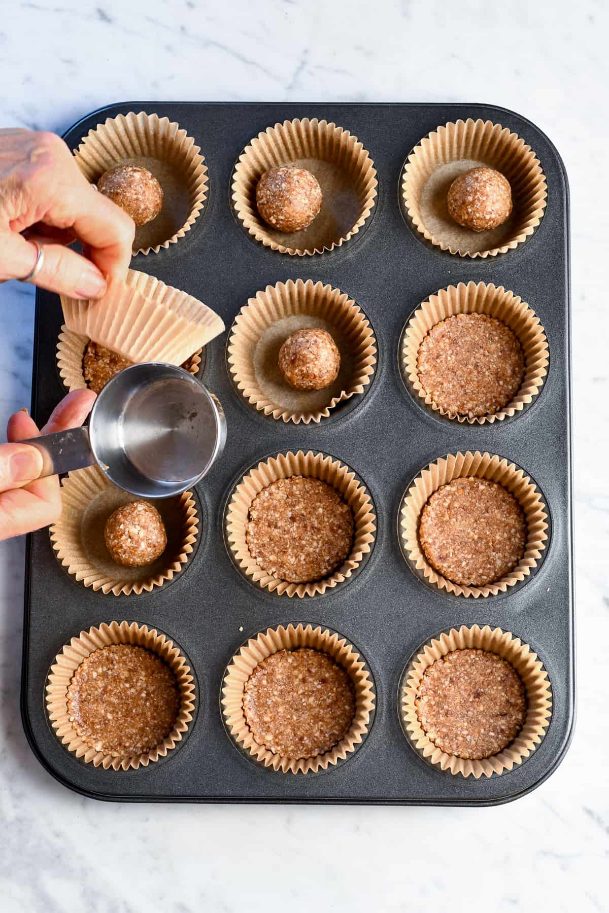 Almond coconut crust in cupcake pan with hands showing how to press the crust into the cupcake liners