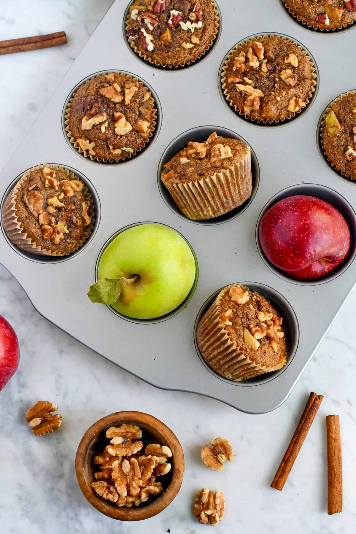 Healthy Apple Muffins baked muffins in muffin tin with a few apples in the muffin tin walnuts and cinnamon sticks on the side